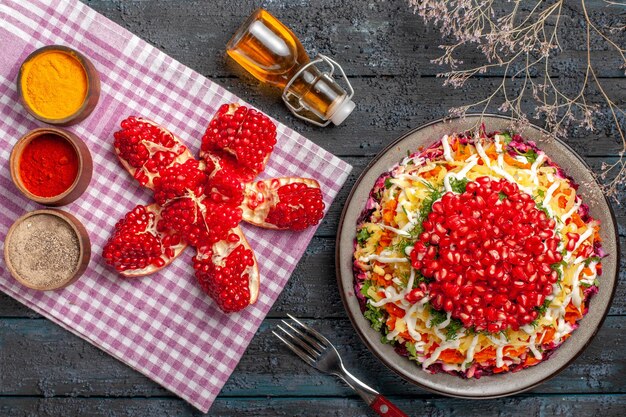 Top view dish and tablecloth dish of pomegranate carrot potato next to the bottle of oil fork tree branches and pilled pomegranate spices on pink-white checkered tablecloth