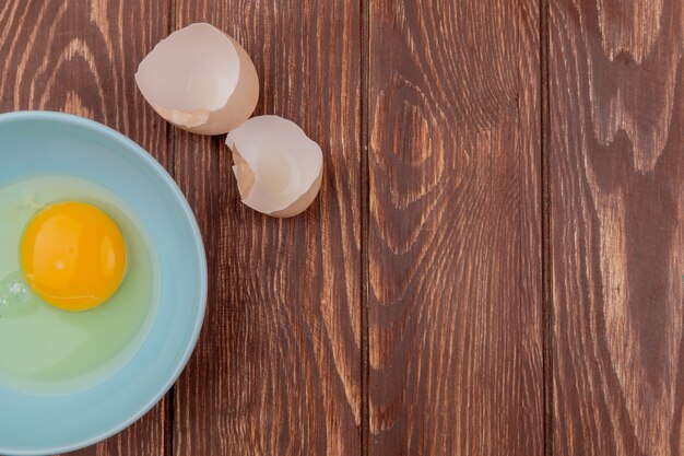 Top view of egg yolk and white on a white bowl with cracked egg shells on a wooden background with copy space