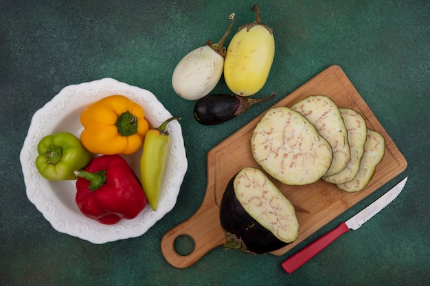 Top view eggplant slices on a cutting board with a knife with bell peppers on a plate on a green background