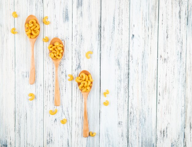 Top view elbow macaroni pasta on spoons on the left with copy space on white wooden background