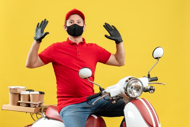 Top view of emotional young adult wearing red blouse and hat gloves in medical mask delivering order sitting on scooter feeling surprised