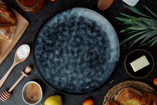 Top view of empty plate with jam lemon kumquats croissants flour butter pineapple leaves on black background