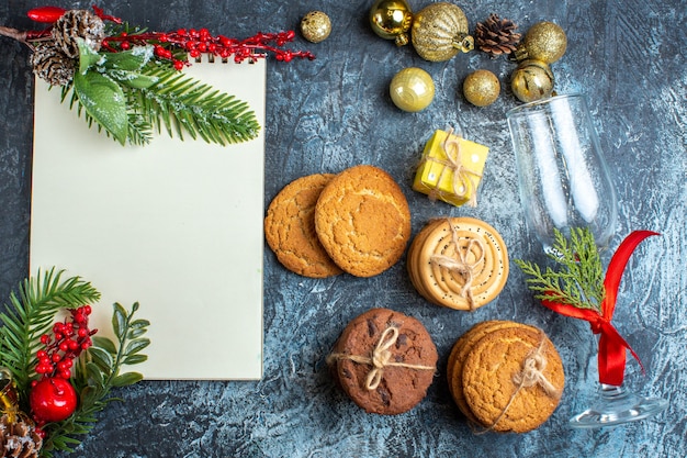 Top view of fallen glass goblet with red ribbon and decoration accessories next to stacked cookies notebook on dark background