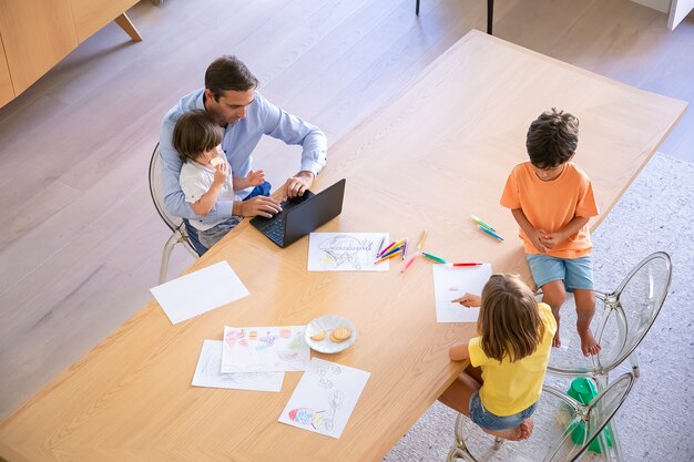 Top view of father with kids sitting at table. Brother and sister drawing doodles with markers. Middle-aged dad working on laptop and holding little son. Childhood, weekend and family time concept