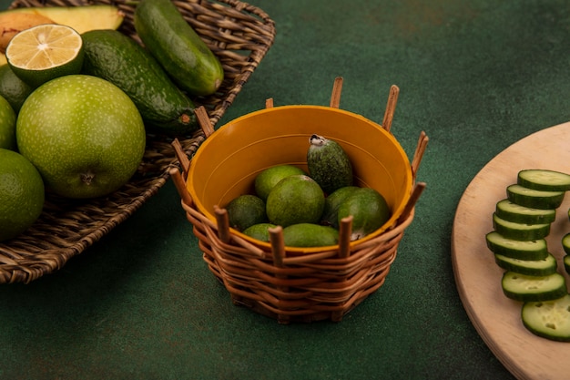 Top view of feijoas on a bucket with chopped slices of cucumber on a wooden kitchen board with green apples avocados cucumber on a wicker tray on a green background