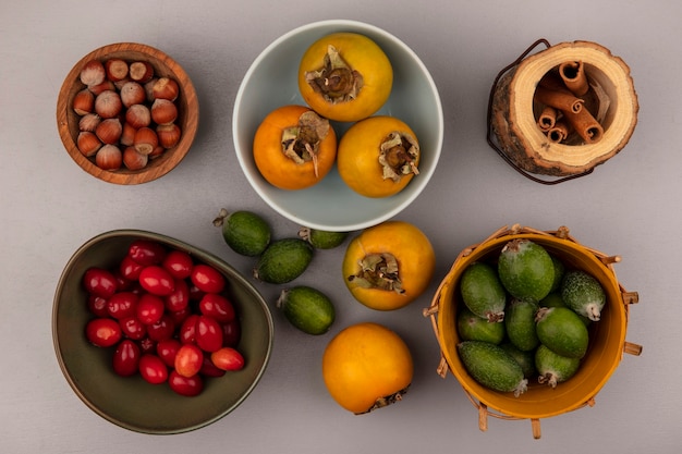 Free photo top view of feijoas on a bucket with persimmons on a bowl with cornelian cherries on a bowl with hazelnuts on a wooden bowl with cinnamon sticks on a grey wall