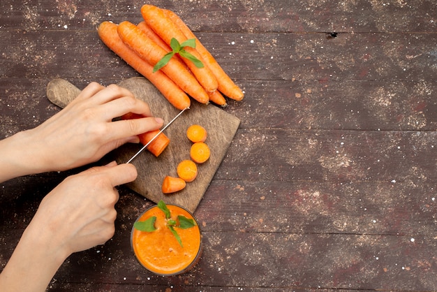 Top view female cutting carrot on brown wooden desk along with fresh carrot juice on brown