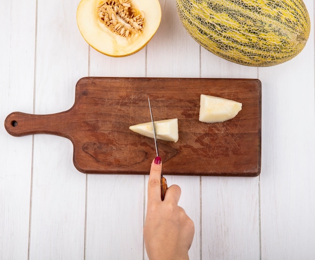 Free photo top view of female hand cutting melon into slices with knife on wooden kitchen board on white wood