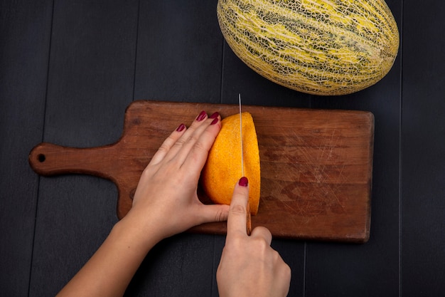 Free photo top view of female hand cutting yellow melon on wooden kitchen board on black