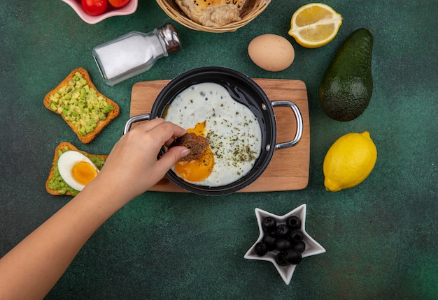 Free Photo top view of female hand holding bread slice with fried egg in a frying pan on wooden kitchen board with black olives salt shaker lemon toast bread with avocado pulp on gre