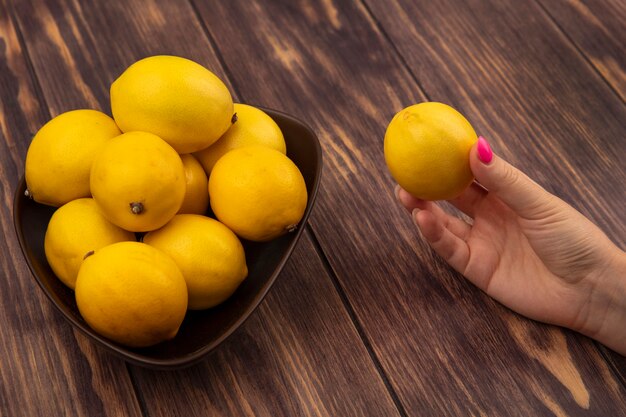 Top view of female hand holding a fresh lemon with a bowl of lemons on a wooden wall