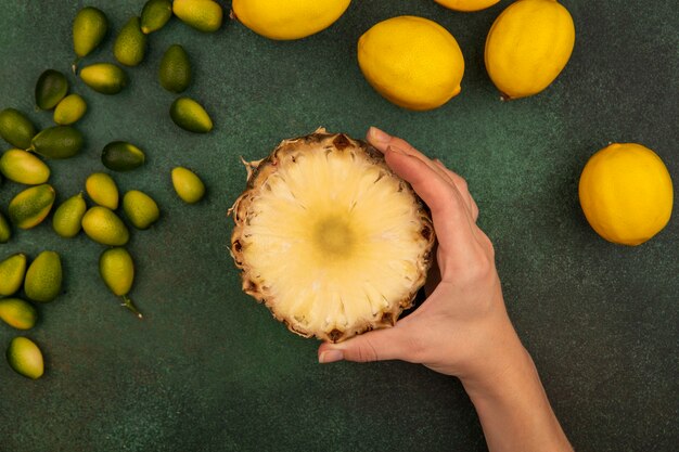 Top view of female hand holding sweet half pineapple with lemons and kinkans isolated on a green wall