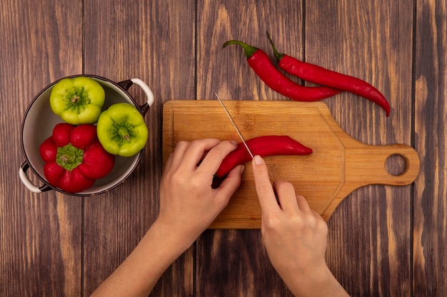 Top view of female hands cutting a chili pepper on a wooden kitchen board with knife on a wooden wall