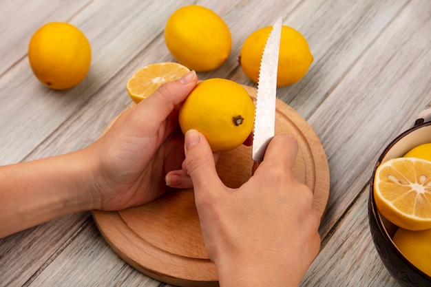 Top view of female hands cutting fresh lemon on a wooden kitchen board with knife with lemons on a bowl with lemons isolated on a grey wooden background