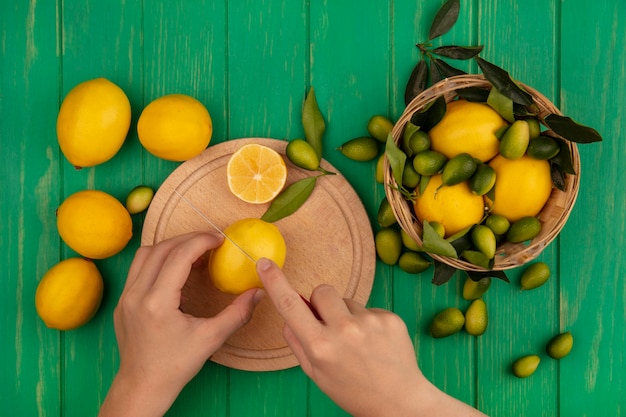 Top view of female hands cutting fresh lemon on a wooden kitchen board with knife with lemons on a bucket on a green wooden wall