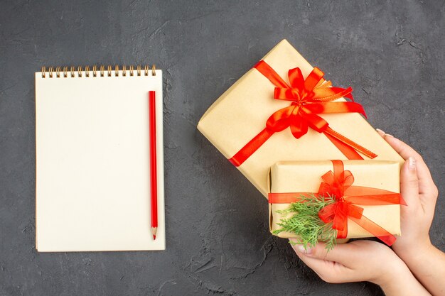 Top view female hands holding big and small xmas gifts in brown paper tied with red ribbon notebook pencil on dark surface