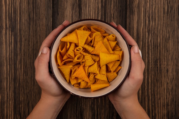 Free photo top view of female hands holding a bowl of cone shape corn snacks on a wooden table