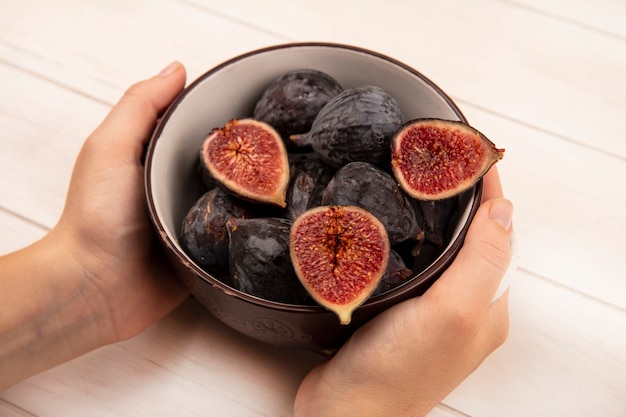 Free photo top view of female hands holding a bowl of fresh sweet black mission figs on a white wooden wall