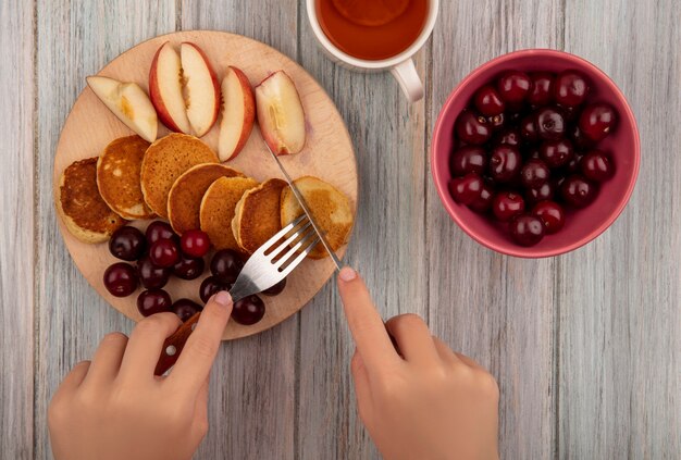 Top view of female hands holding fork and knife with pancakes and cherries peach slices on cutting board with bowl of cherry and tea on wooden background