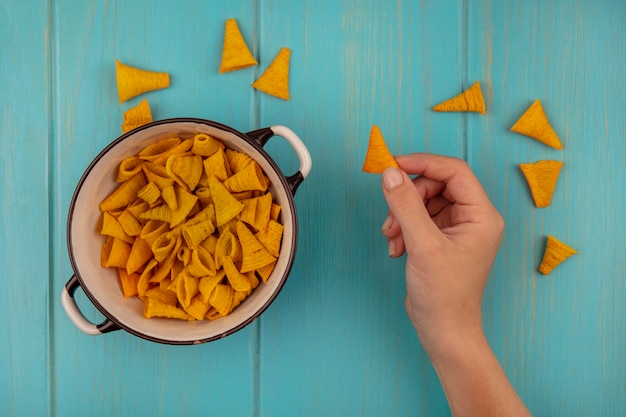 Top view of female hands holding tasty cone shape corn snacks with a bowl of corn snacks on a blue wooden table