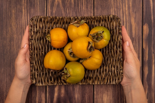 Free Photo top view of female hands holding a wicker tray of fresh persimmon fruits on a wooden table