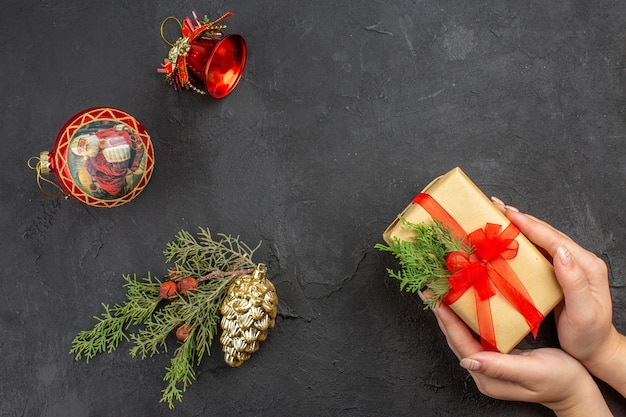 Top view female hands holding xmas gift in brown paper tied with red ribbon xmas tree ornaments on dark background