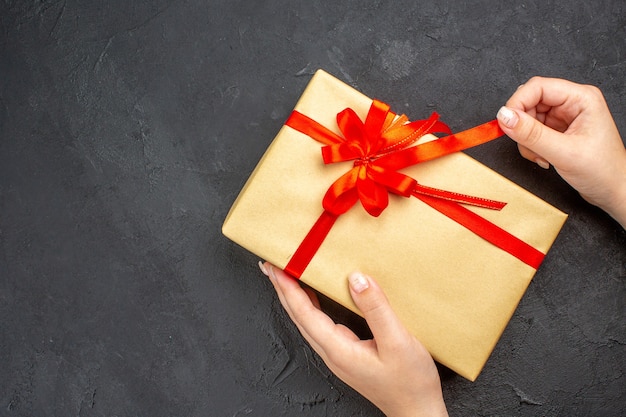 Top view female hands opening xmas gift in brown paper with red ribbon on dark background free space