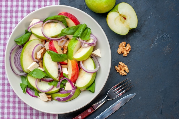 Top view fresh apple salad on round plate green apples walnut purple and white checkered tablecloth fork and knife on dark table