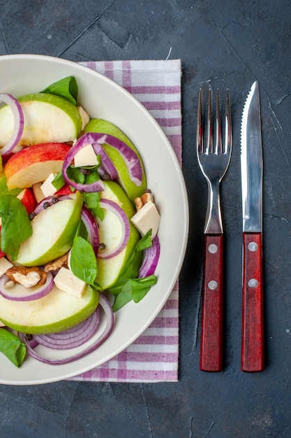 Free photo top view fresh apple salad on round plate on purple and white checkered tablecloth knife and fork on black background