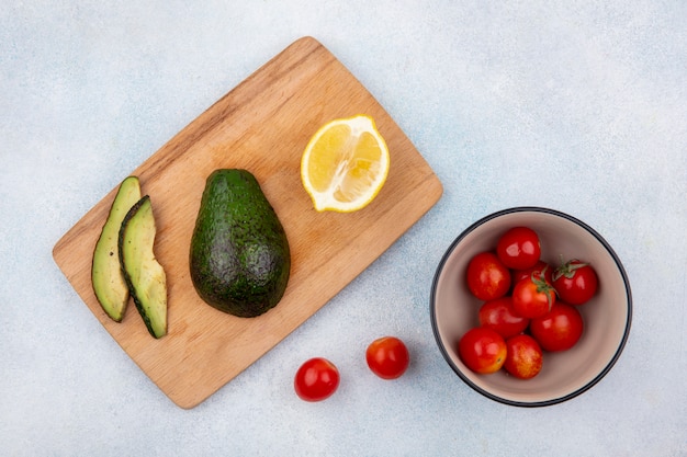 Top view of fresh avocado on wood kitchen board with lemonnd tomatoes on bowl on white