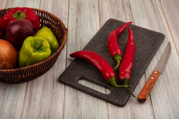 Top view of fresh bell peppers on a bucket with onions with chili peppers on a black kitchen board with knife on a grey wooden background