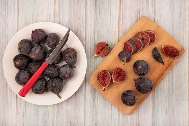 Top view of fresh black figs on a white bowl with knife with halved figs on a wooden kitchen board on a grey wooden wall