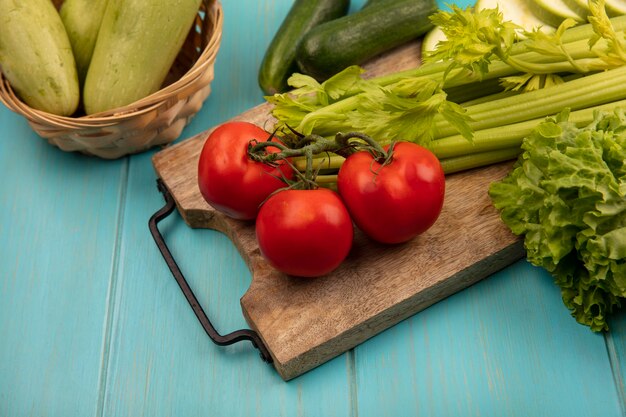 Top view of fresh celery on a wooden kitchen board with tomatoes lettuce with zucchinis on a bucket on a blue wooden surface