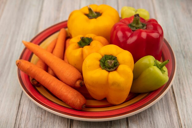 Top view of fresh colorful bell peppers on a plate with carrots on a grey wooden surface