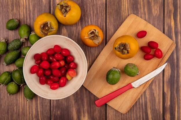 Top view of fresh cornelian cherries on a bowl with persimmon and feijoas on a wooden kitchen board with knife on a wooden surface