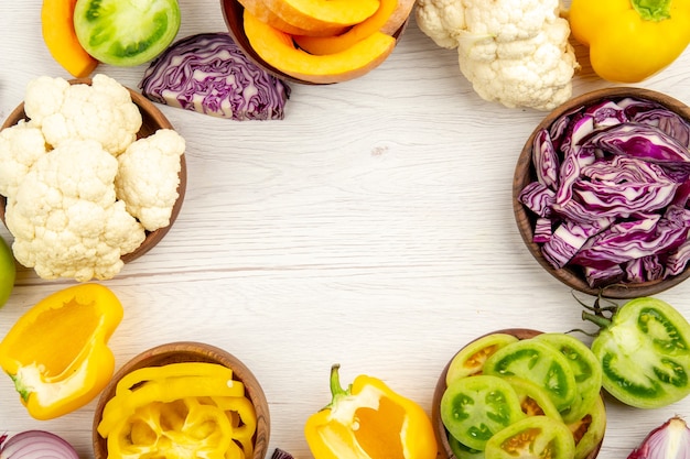 Free Photo top view fresh cut vegetables green tomatoes red cabbage onion pumpkin cauliflower yellow bell pepper in bowls on white wooden surface free space in center