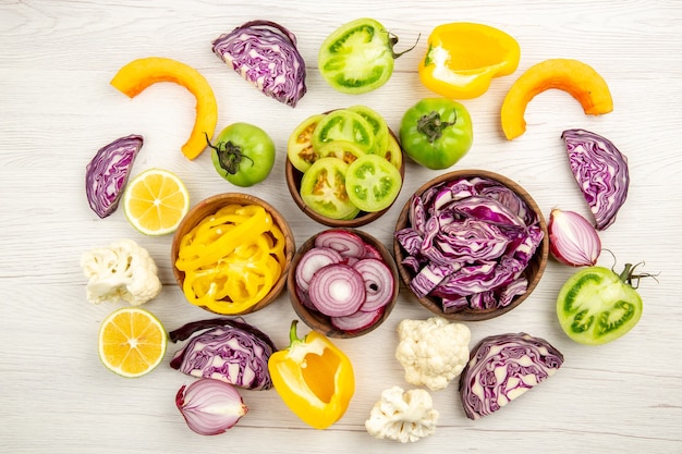 Top view fresh cut vegetables red cabbage green tomato pumpkin red onion yellow bell pepper caulifower lemon in small bowls on white wooden surface