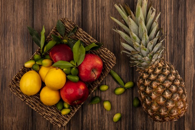 Top view of fresh fruits such as apples lemons and kinkans on a wicker tray with pineapple isolated on a wooden wall