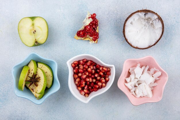 Top view of fresh fruits such as half green apple with chopped apple slices with pomegranate seeds in a white bowl and coconut with pulps of coconut in a pink bowl on white