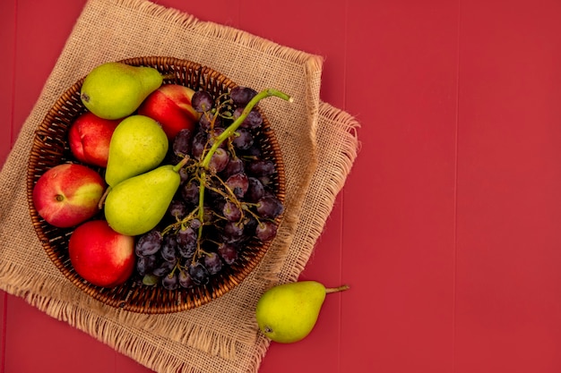 Top view of fresh fruits such as pearpeach grape on a wooden bowl on a sack cloth on a red background with copy space