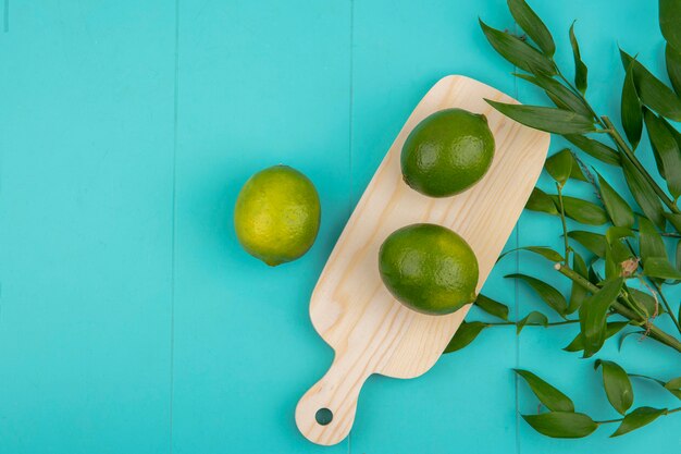 Top view of fresh green lemons on wood kitchen board with leaves on blue with copy space