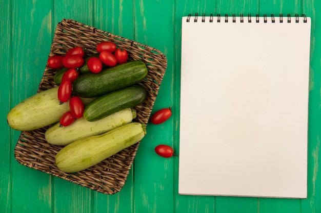 Free photo top view of fresh and healthy vegetables such as plum tomatoes cucumbers and zucchinis on a wicker tray on a green wooden wall with copy space