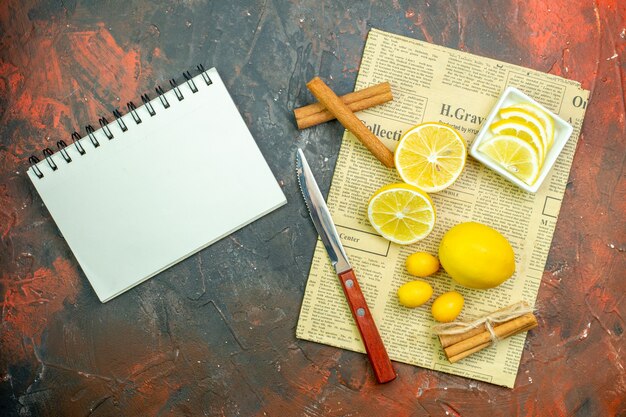 Top view fresh lemons cumcuats cinnamons lemon slices in small bowl knife on newspaper notebook on dark red table