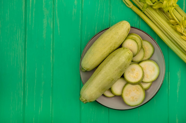 Top view of fresh light green zucchinis on a plate with celery isolated on a green wooden wall with copy space