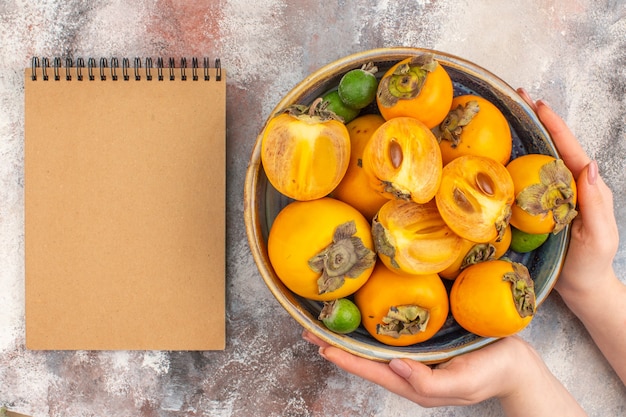 Free photo top view fresh persimmons feykhoas in a bowl in female hand a notebook on nude background