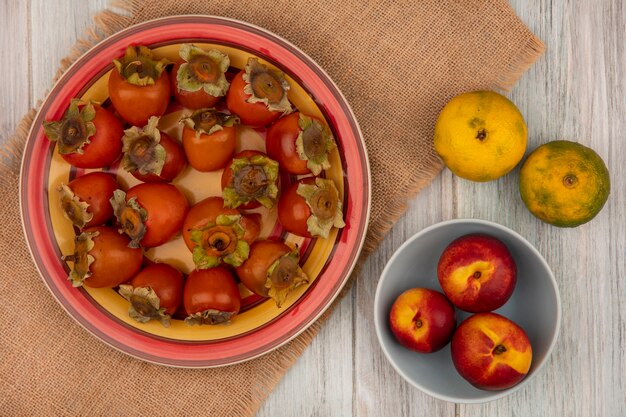 Top view of fresh persimmons on a plate on a sack cloth with peaches on a bowl with tangerines isolated on a grey wooden wall