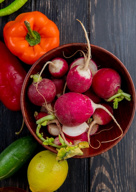 Free photo top view of fresh radish in a wood bowl and fresh colorful bell peppers cucumbers and lemon on dark wood