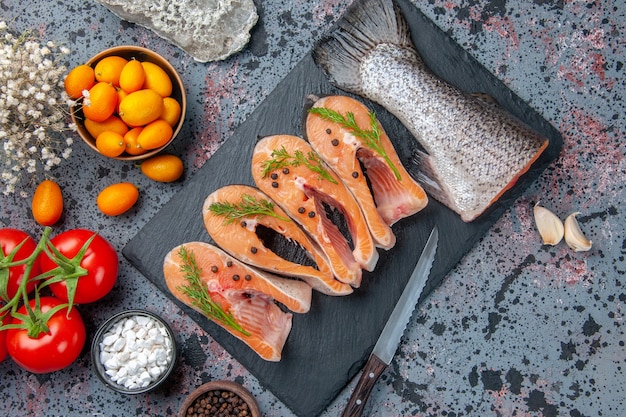 Top view of fresh raw fishes greens on dark color tray spices tomatoes with stems garlics flowers knife kumquats on blue black mix colors table