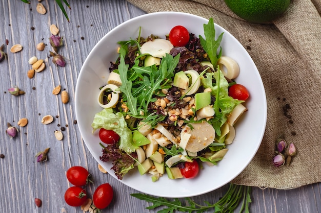 Top view of fresh salad with cheese cherry tomatoes avocado and roasted peanuts in a white bowl on rustic background