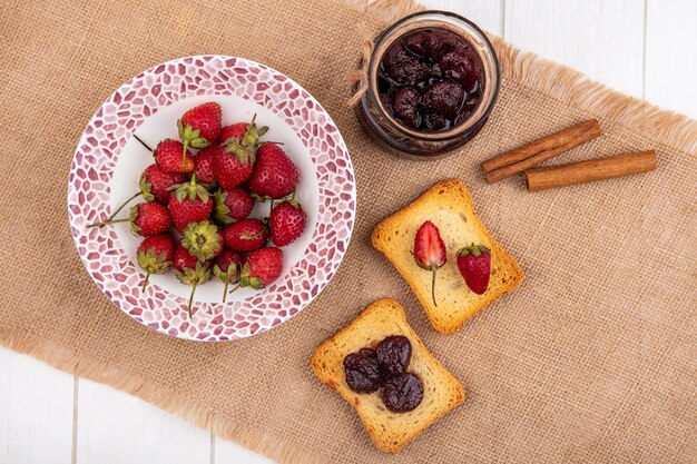 Top view of fresh strawberries on a bowl on a sack cloth with strawberry jam with cinnamon sticks on a white wooden background